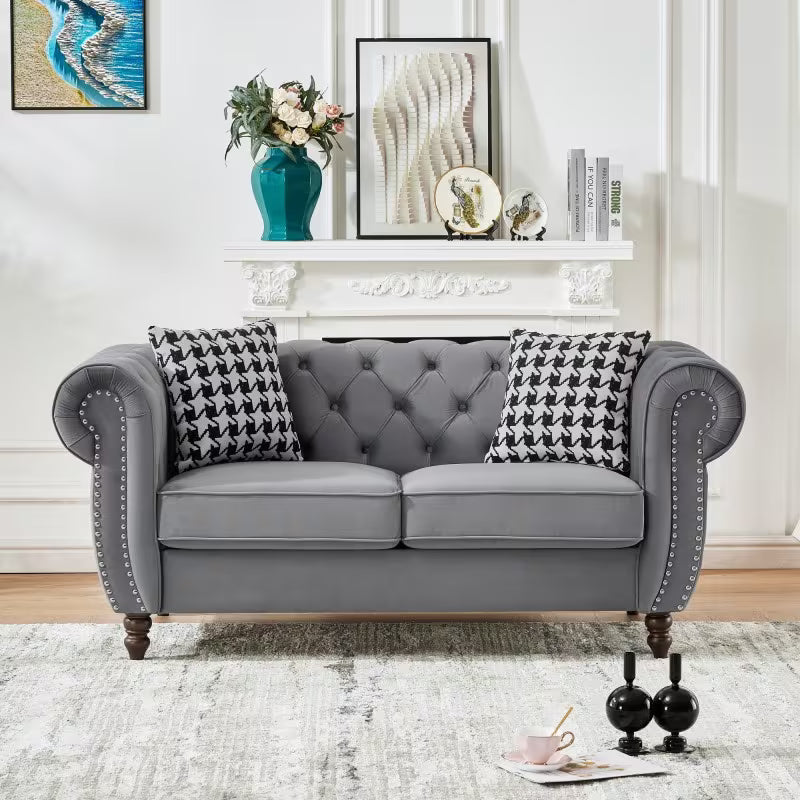 A gray tufted loveseat with rolled arms, nailhead trim, and dark wooden legs, styled in a living room setting with two black-and-white houndstooth pillows, a white mantel, and framed wall art in the background.